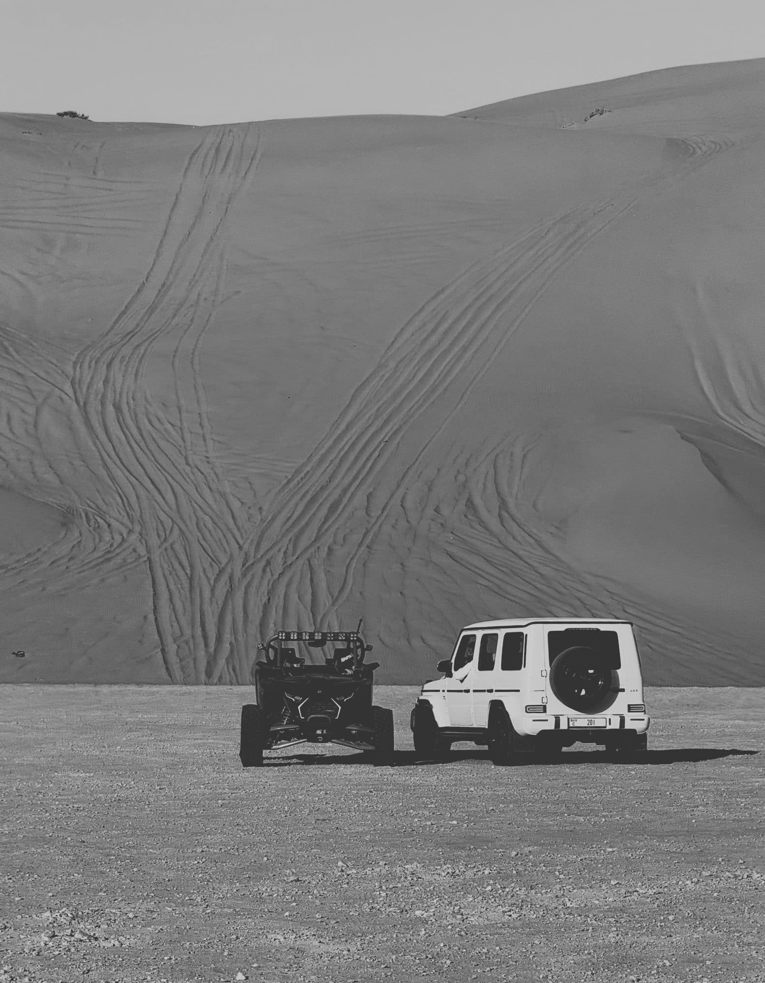 A white G-Class and a dune buggy parked at the foot of a large dune, in black and white.
